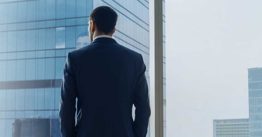 The back of a man dressed in a suit looking out a window at another sky scraper.
