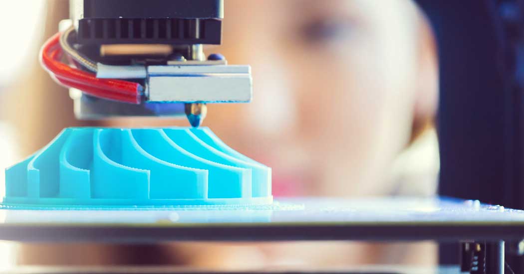 A 3d printer printing a blue piece with a blonde woman watching in the background.