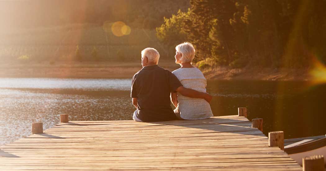 An older man with his arm around an older woman, sitting at the end of a dock watching the sun set.