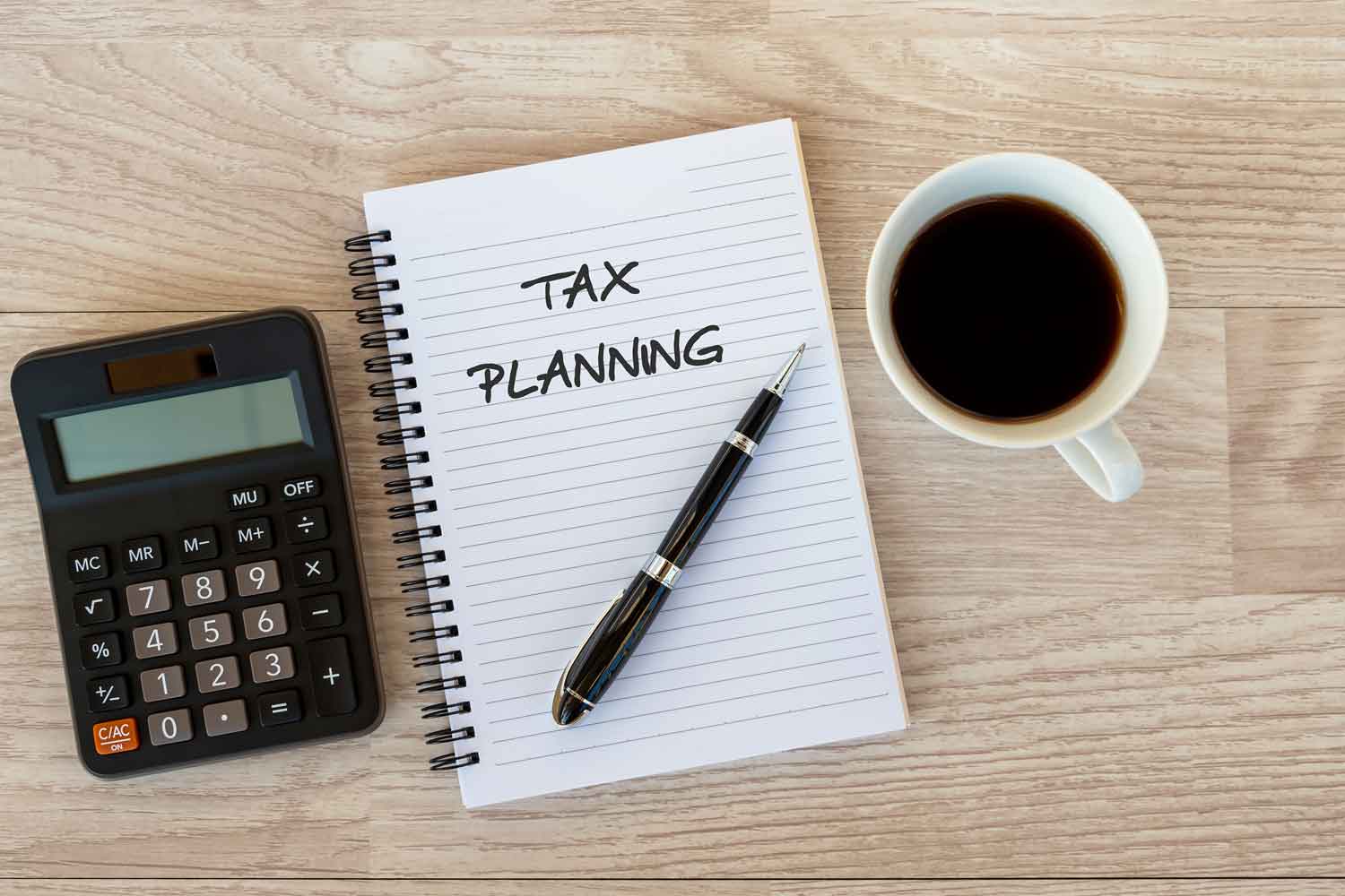 11Notebook with the words "Tax Planning" on it with a pen, a cup of coffee and a calculator on a desk.